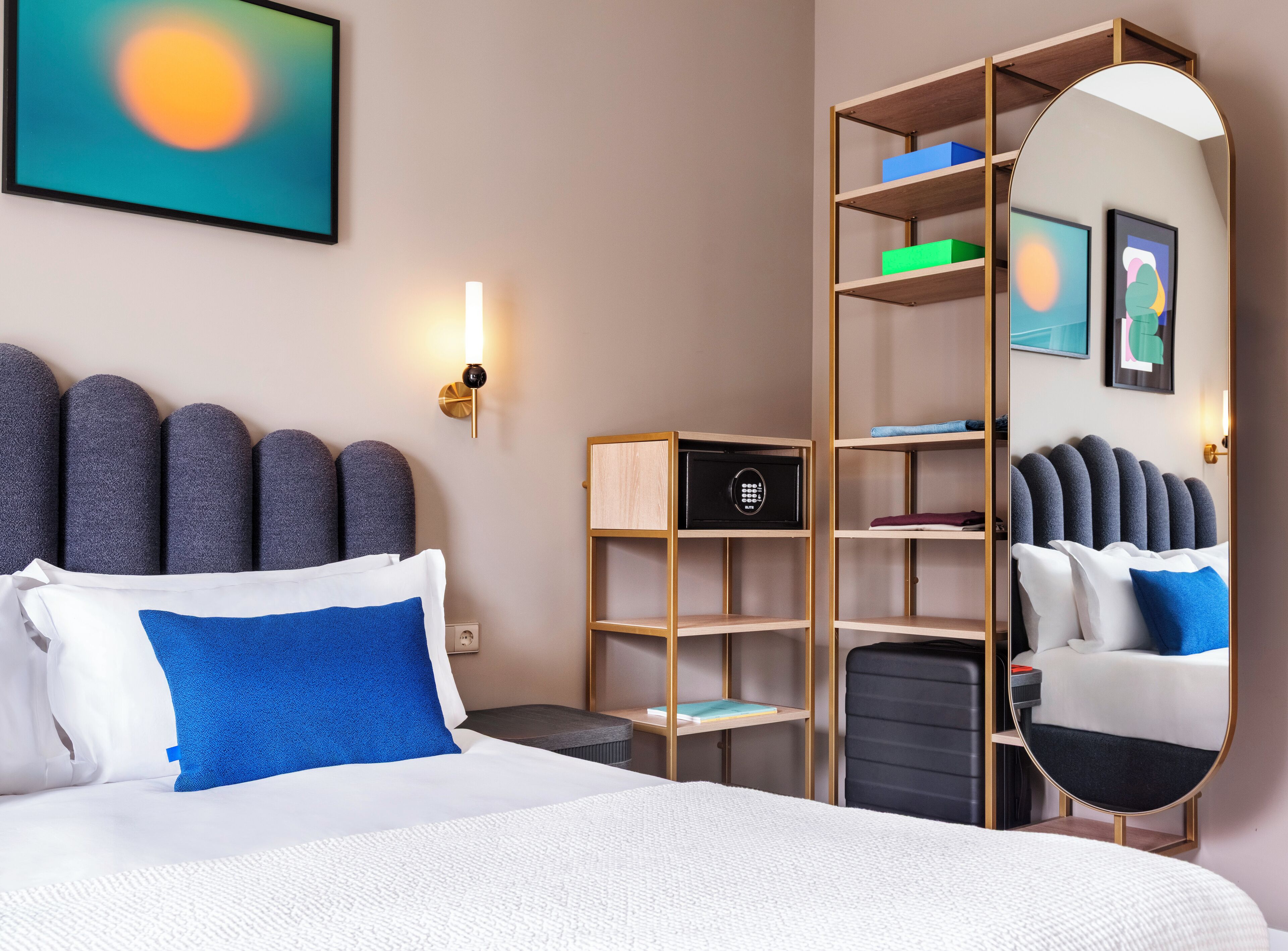 A modern hotel room with a large bed, a bedside table, a mirror, a wardrobe, and a wall-mounted artwork.