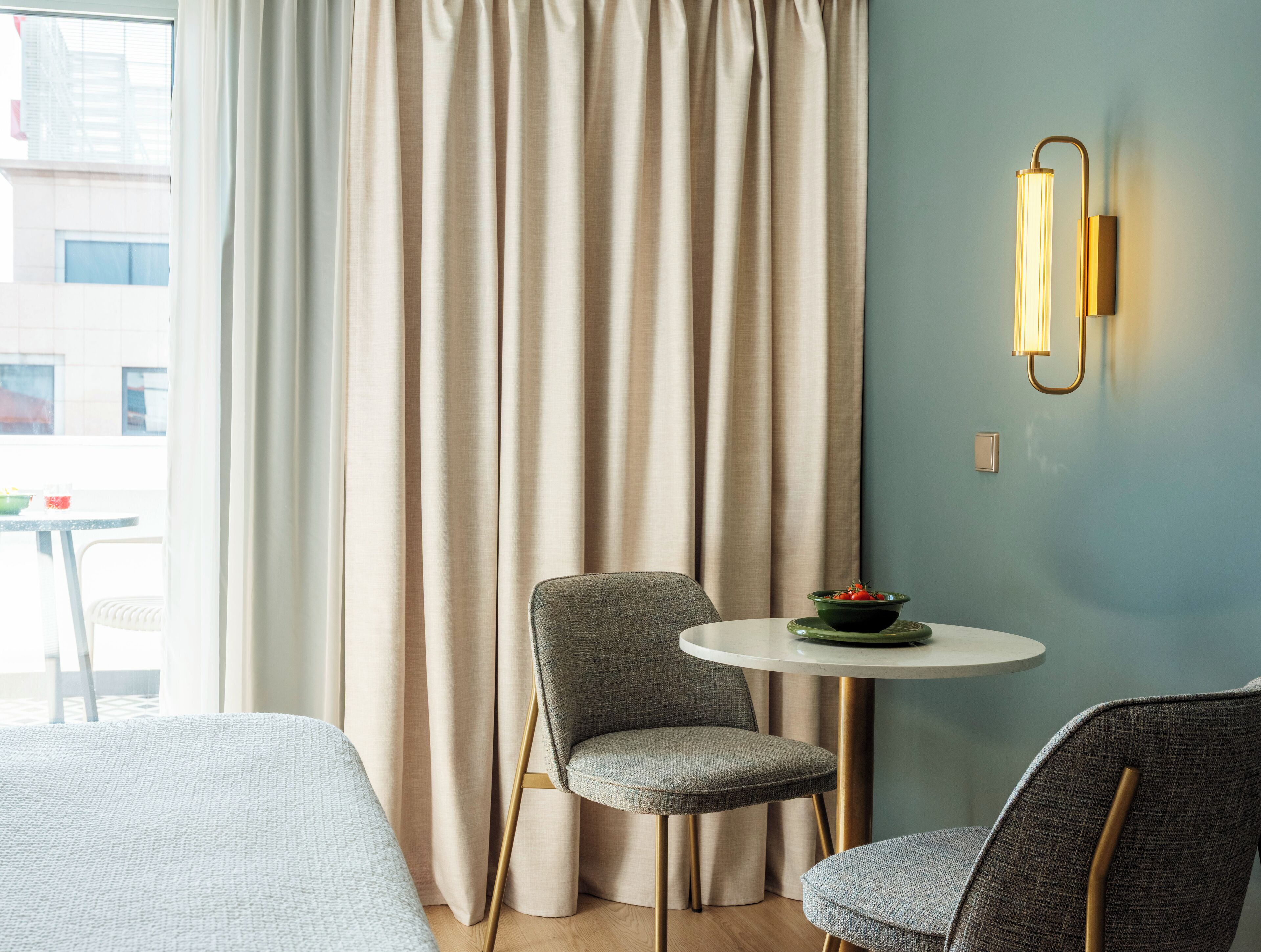 Une chambre d’hôtel avec un lit, une table ronde sur laquelle se trouve un saladier rempli de fruits, une chaise et un luminaire fixé au mur.