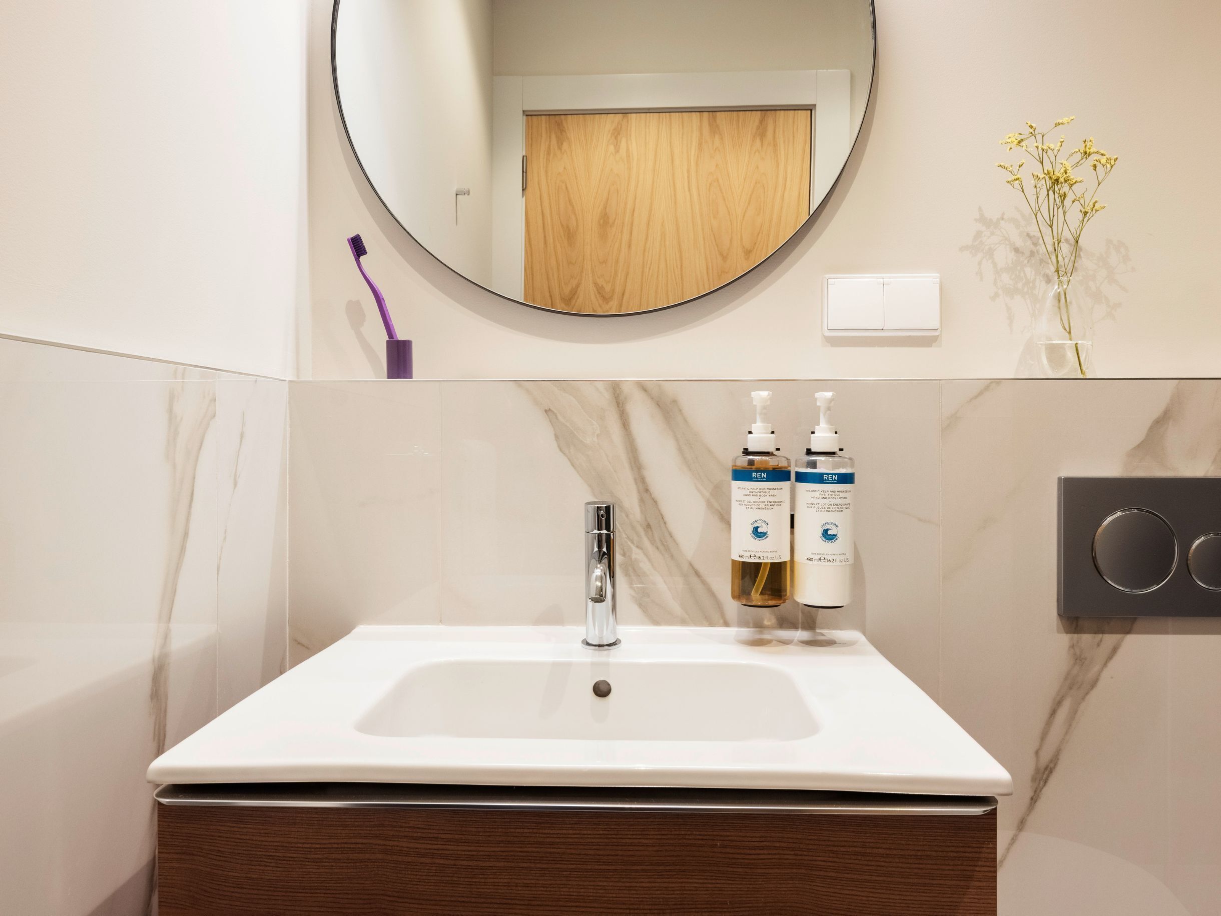 Un baño con un lavabo blanco, un espejo, una puerta de madera y dos botellas de crema corporal REN.