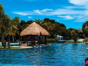 A swimming pool with a thatched-roof structure, palm trees, and a clear blue sky. - ILOA RESIDENCE AP 103 A (Barra de São Miguel)