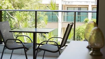 Un balcón con una mesa y sillas, barandilla de vidrio y vistas a edificios y vegetación.