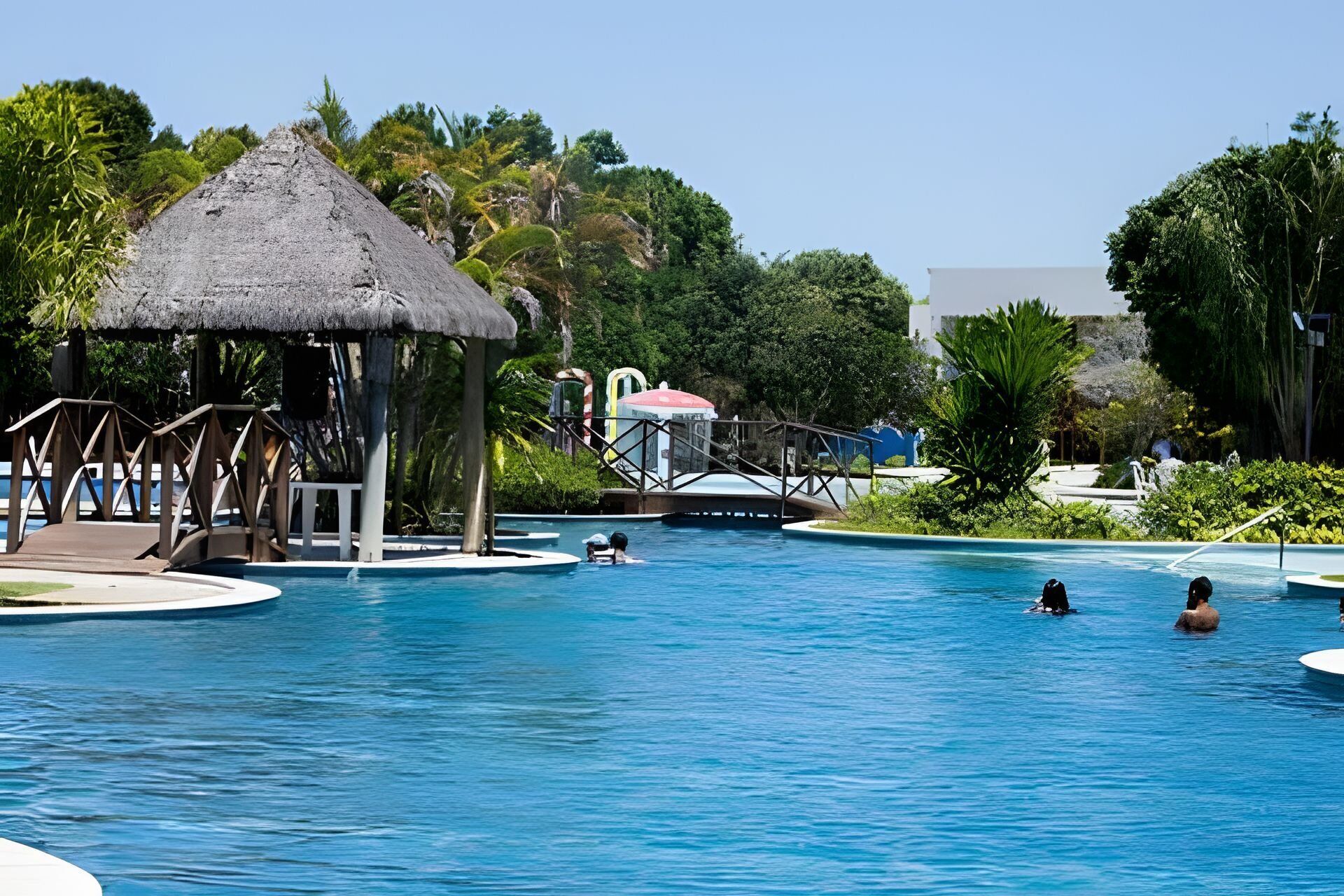A swimming pool with a thatched-roof pavilion, palm trees, and a bridge.