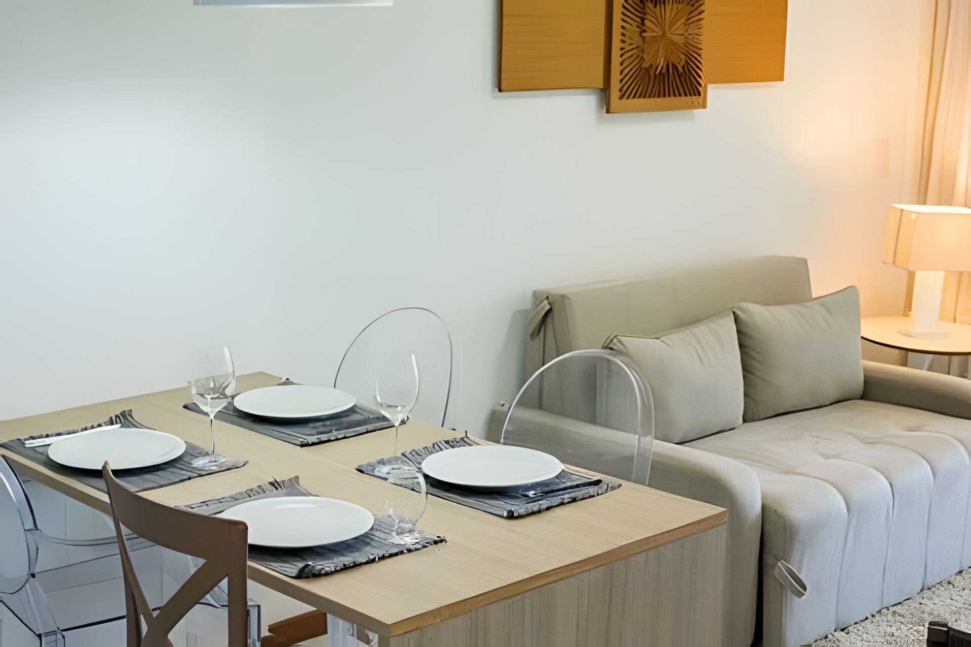 Uma sala de jantar com uma mesa de madeira, quatro pratos brancos e copos de vidro transparente. Um sofá bege e um abajur estão ao fundo.