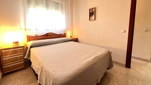 Ein Schlafzimmer mit Bett, Nachttischlampen, einem Nachttisch, einem Fenster mit leichtem Vorhang und einem gerahmten Bild an der Wand.