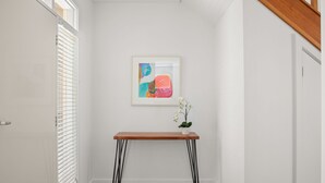 Um corredor com porta branca, parede branca, quadro abstrato emoldurado, mesa de madeira com pés pretos e uma orquídea em vaso.