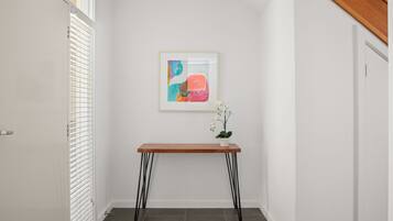 A hallway with a white door, a white wall, a framed abstract painting, a wooden table with black legs, and a potted orchid.