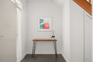 A hallway with a white door, a white wall, a framed abstract painting, a wooden table with black legs, and a potted orchid. - Flora  Pet Friendly Coastal Hampton Retreat (Hampton)