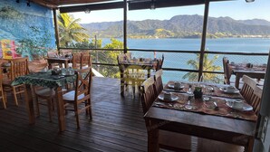 A restaurant with wooden tables and chairs, a mural on the wall, and a view of a lake and mountains. - Residencial Solariun Ilhabela (Ilhabela)