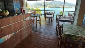 A restaurant with wooden flooring, tables and chairs, a bar area with a bowl of fruit, and a view of the sea and mountains. - Residencial Solariun Ilhabela (Ilhabela)