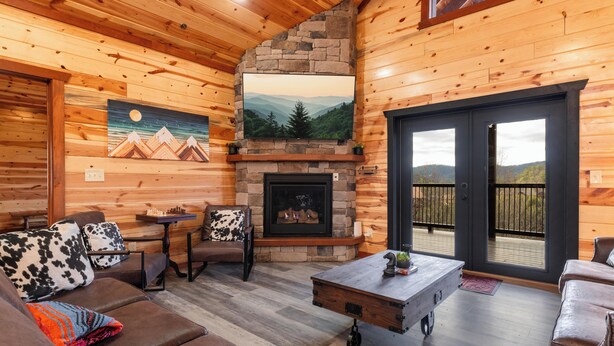 L’intérieur chaleureux d’un chalet avec une cheminée en pierre, des murs en bois et une vue sur les montagnes, visible à travers de grandes portes coulissantes.