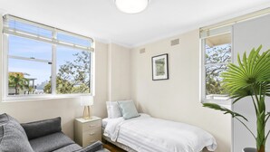 Quarto com cama, sofá cinza, mesa de cabeceira com abajur, janela ampla e planta em vaso.