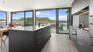 Une cuisine moderne dotée d’un grand îlot central, de meubles de rangement aux couleurs sombres et de grandes fenêtres offrant une vue sur le paysage urbain et les collines.