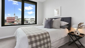 Une chambre avec une grande fenêtre, un lit recouvert d’un plaid à carreaux, une table de chevet avec une lampe et un tableau encadré accroché au mur.