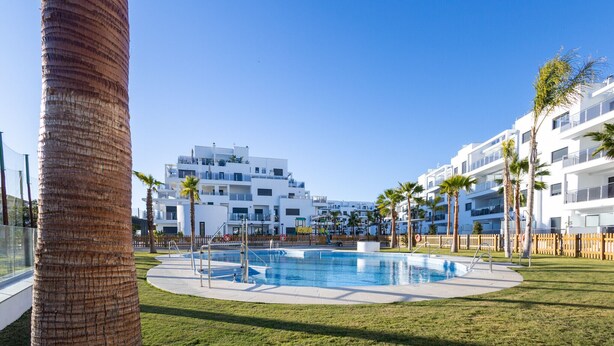 Una piscina rodeada de césped, palmeras, edificios blancos modernos al fondo y un cielo azul despejado.