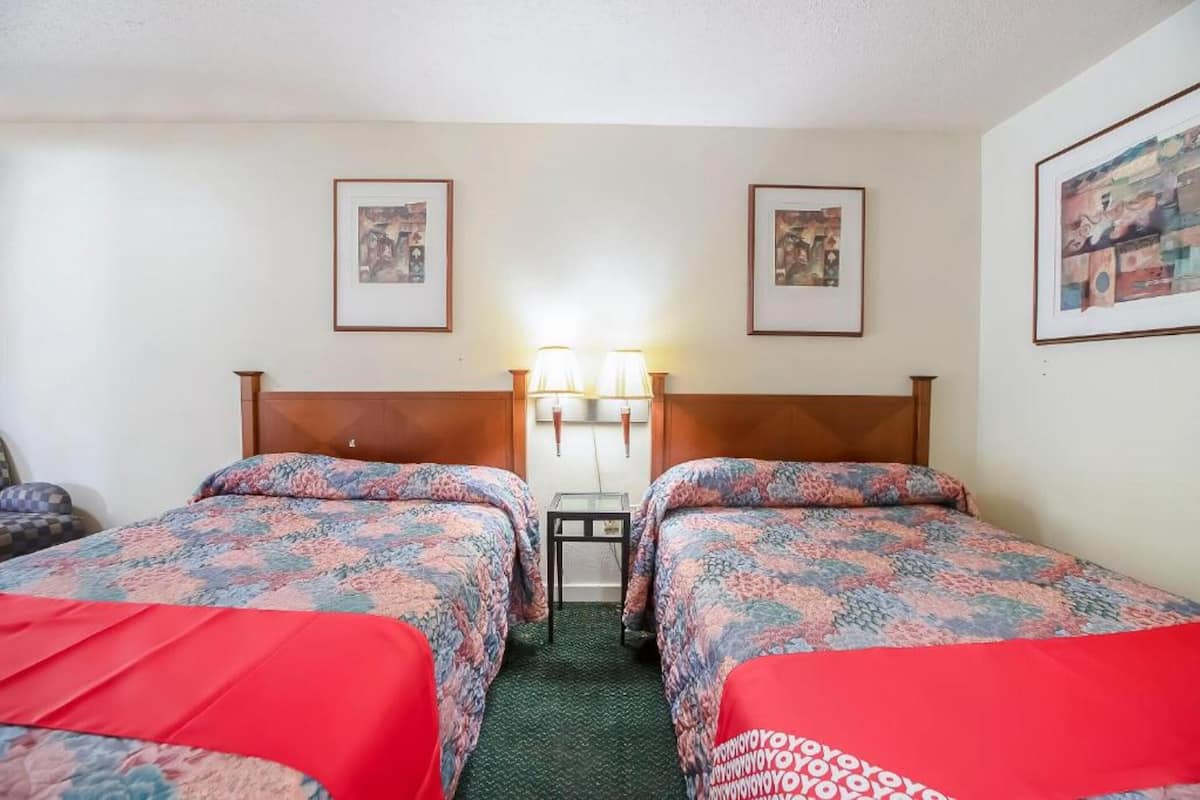 A hotel room with two beds, each with a floral-patterned bedspread and red pillows. There are framed pictures on the walls and a small table between the beds.