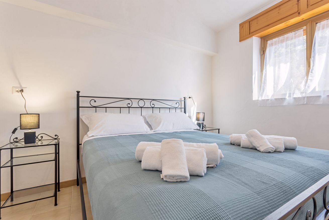 Una cama bien hecha con toallas enrolladas, una lámpara de noche y una ventana con cortinas translúcidas.