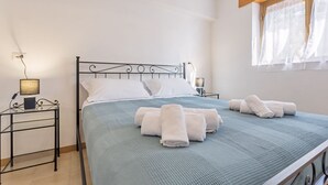 Een net opgemaakt bed met opgerolde handdoeken, een bedlampje en een raam met dunne gordijnen.