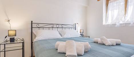 Una cama bien hecha con toallas enrolladas, una lámpara de noche y una ventana con cortinas translúcidas.