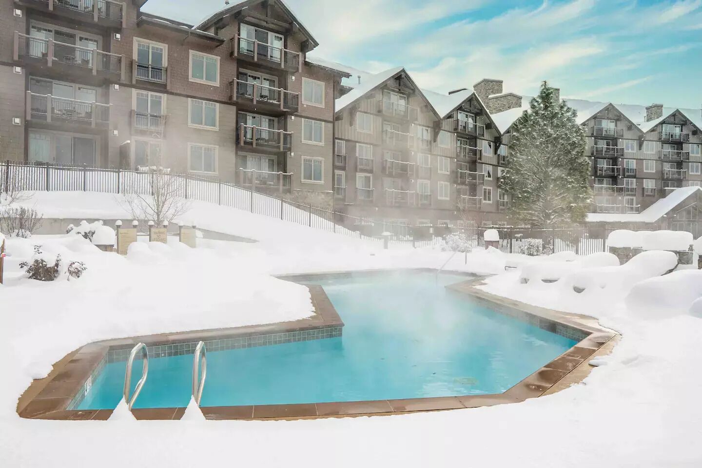 Un espace extérieur avec une piscine entourée de neige, l’eau de la piscine étant d’un bleu limpide, et l’ensemble étant bordé d’immeubles à plusieurs étages dotés de balcons.