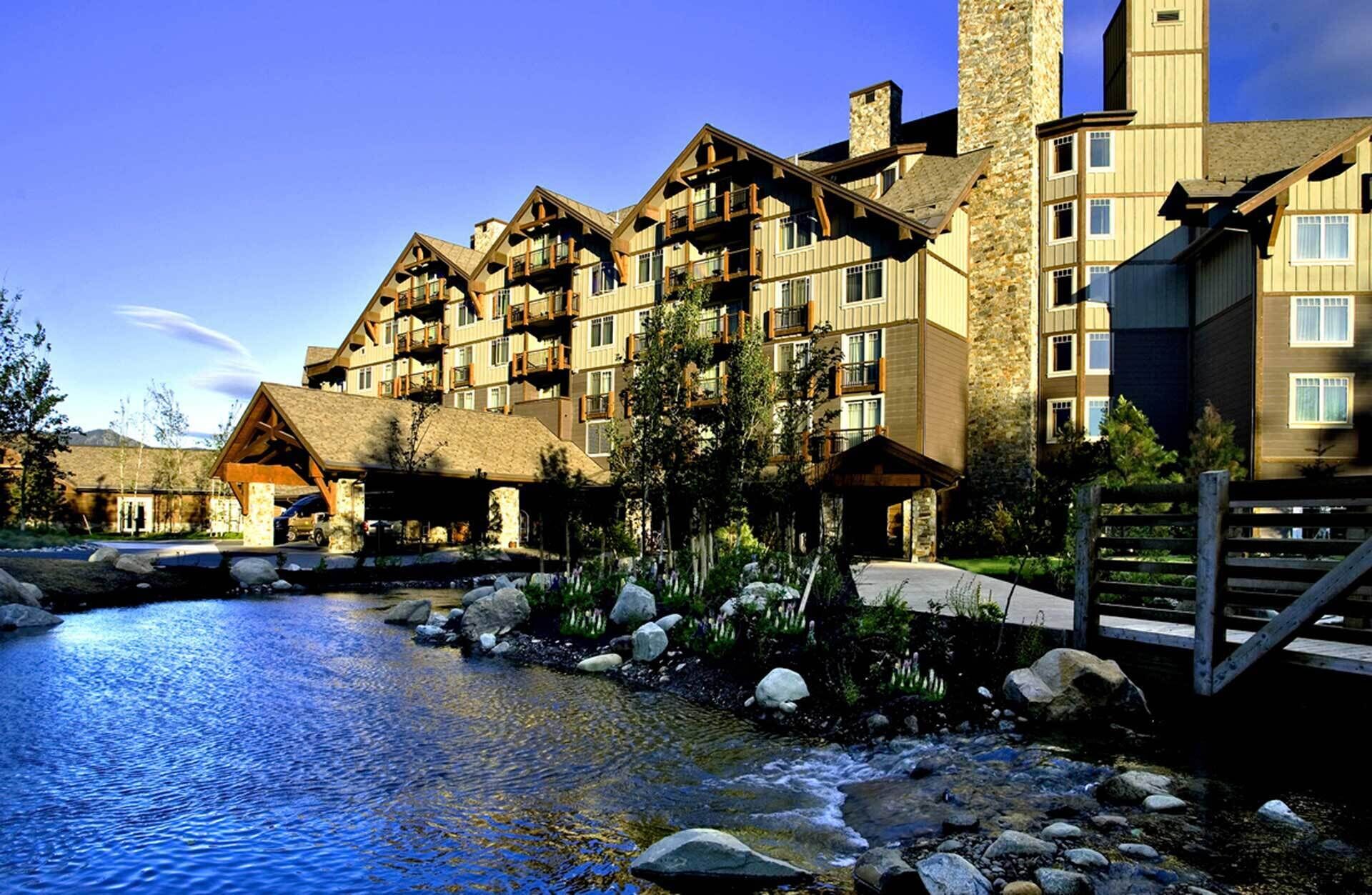 Ein mehrgeschossiges Hotel mit einer Steinfassade und hölzernen Balkonen, ein Poolbereich in einer natürlichen Landschaft und eine Brücke zum Eingang.