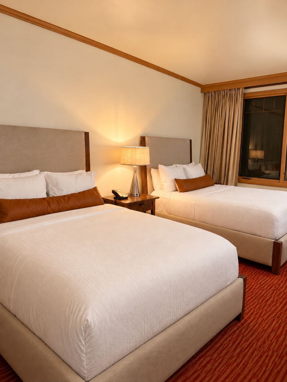 A hotel room with two beds, a nightstand with a lamp, and a large window with curtains.