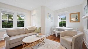 Amplio salón con un sofá color beige, una mesa de centro redonda y un sillón blanco. Se observan dos ventanas con marcos blancos, una chimenea blanca y una pequeña mesa con sillas.