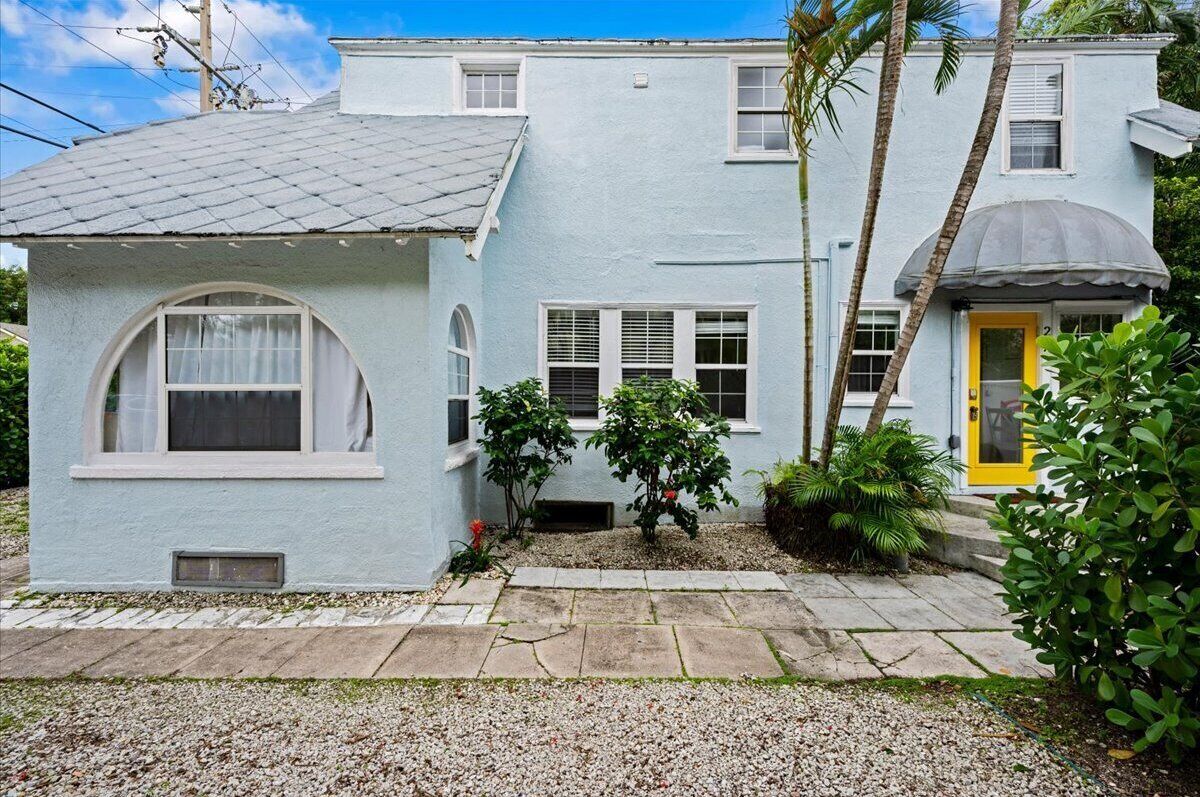 Una casa de dos pisos con fachada azul claro, una puerta amarilla y un pequeño jardín con palmeras y arbustos.