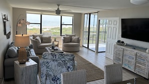 Un salon spacieux dotĂ© dâune grande fenĂȘtre donnant sur la plage, dâun ventilateur de plafond et dâun tĂ©lĂ©viseur Ă Ă©cran plat fixĂ© au mur.
