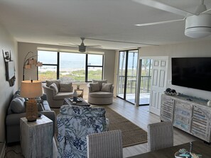 Ein großzügiges Wohnzimmer mit einem großen Fenster und Meerblick, einem Deckenventilator und einem an der Wand befestigten Flachbildfernseher.