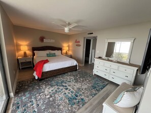 Une chambre à coucher comprenant un lit, des tables de chevet, une commode, un ventilateur de plafond, un tapis et une porte donnant sur une autre pièce.