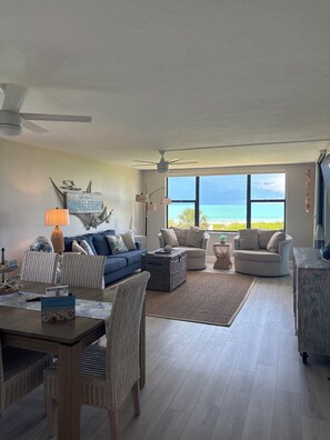 Ein geräumiges Wohnzimmer mit einem großen Fenster mit Meerblick, ausgestattet mit einem blauen Sofa, beigen Sesseln und einem Essbereich.