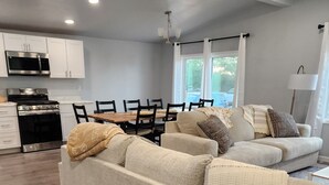 A modern living room with a large beige sectional sofa, a dining area with a wooden table and chairs, a kitchen with white cabinets and stainless steel appliances, and a large window providing natural light. - Red Mesa house  (Page)