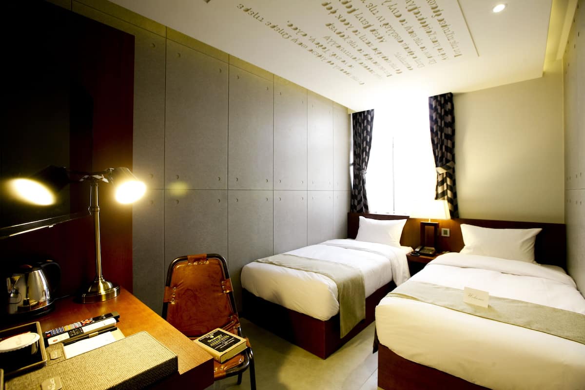 Ein Hotelzimmer mit zwei Betten, einem Schreibtisch mit Lampe, einem Sessel und einem Nachttisch mit Telefon.