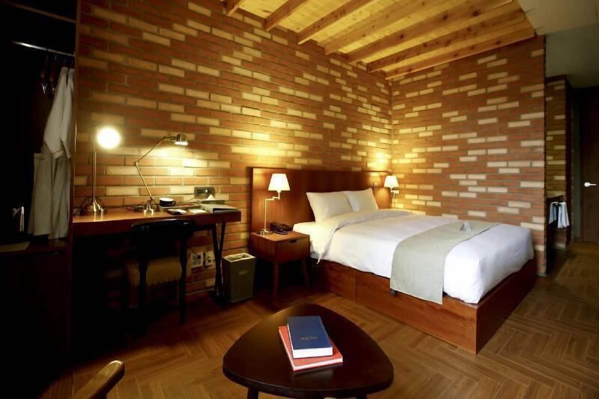 Ein Zimmer mit Holzboden, einer Ziegelwand, einem Bett mit weißer Bettwäsche, einem Schreibtisch mit Lampe und einem kleinen Tisch mit einem Buch.