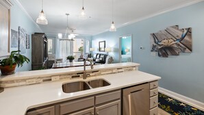 A modern kitchen with a large island, stainless steel appliances, pendant lighting, and a view into the living area.