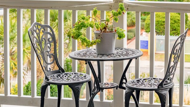 A balcony with two metal chairs and a small round table, overlooking a beach and a pier.