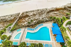 A pool area with two separate pools, surrounded by lounge chairs and umbrellas, with a beach and rocky terrain in the background. - Bluewater 104 | Gulf Front Garden Condo | Indoor, Outdoor & Kiddie Pool | My Beach Getaways (Orange Beach)