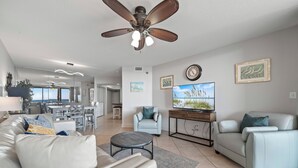 A modern living room with a sofa, armchair, and coffee table, a dining area with chairs, a ceiling fan, and a TV on the wall.