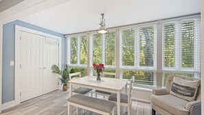 A room with a white table and bench, a beige armchair with a pillow, a potted plant, and a large window with blinds. - Contemporary Seascape Villa, Popular Kiawah West Beach, 1BR, Steps to Beach! (Kiawah Island)