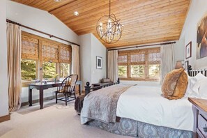 A bedroom with a wooden ceiling, a large bed, a desk, and two windows with curtains.