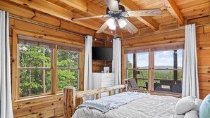 Un dormitorio de cabaña de madera con cama, ventilador de techo y ventanas amplias que dan al bosque.