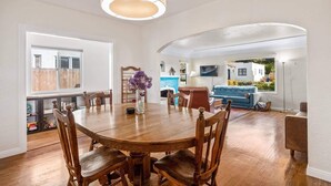 A spacious dining area with a wooden table and chairs, a vase with purple flowers, and a large window with a view of a pool and greenery. - Amazing Reviews, Great Location (Ventura)
