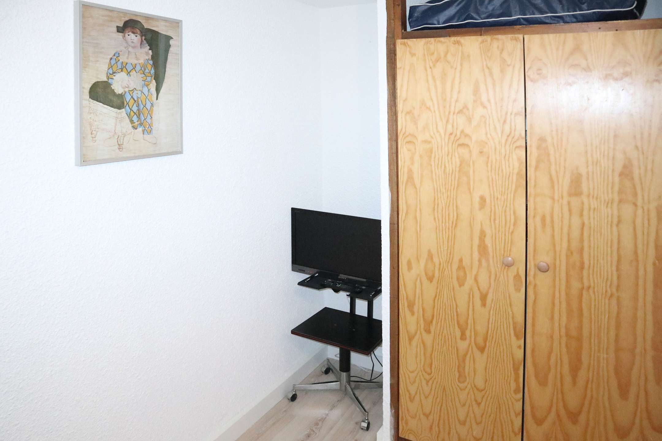 A room with a wooden wardrobe, a television on a stand, and a painting on the wall.