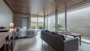 A modern bedroom with a bed, a sofa, a dining table, and large windows overlooking a garden.