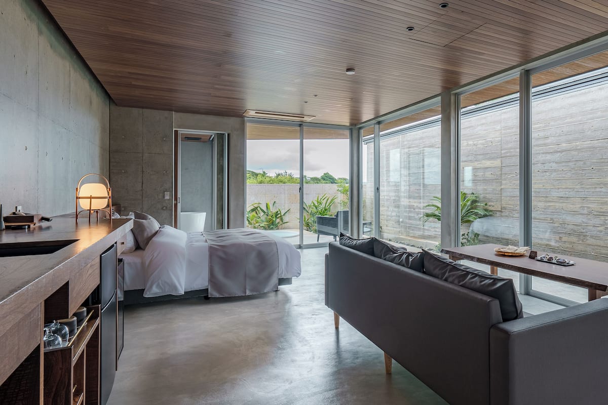 A modern bedroom with a bed, a sofa, a dining table, and large windows overlooking a garden.