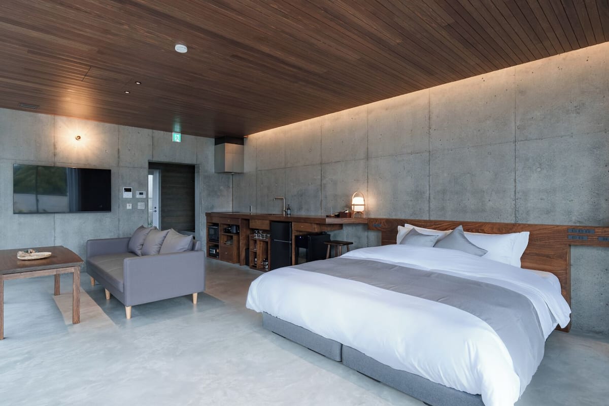 A modern bedroom with a large bed, a sofa, a small table, and a built-in kitchen area.