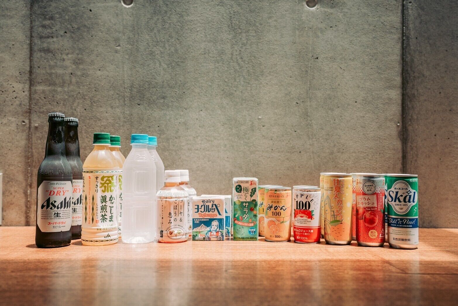 A variety of beverage containers on a wooden surface against a concrete wall.