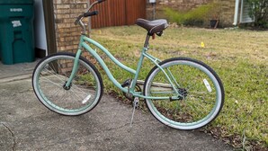 A teal bicycle parked on a concrete surface with grass and a brick wall in the background.