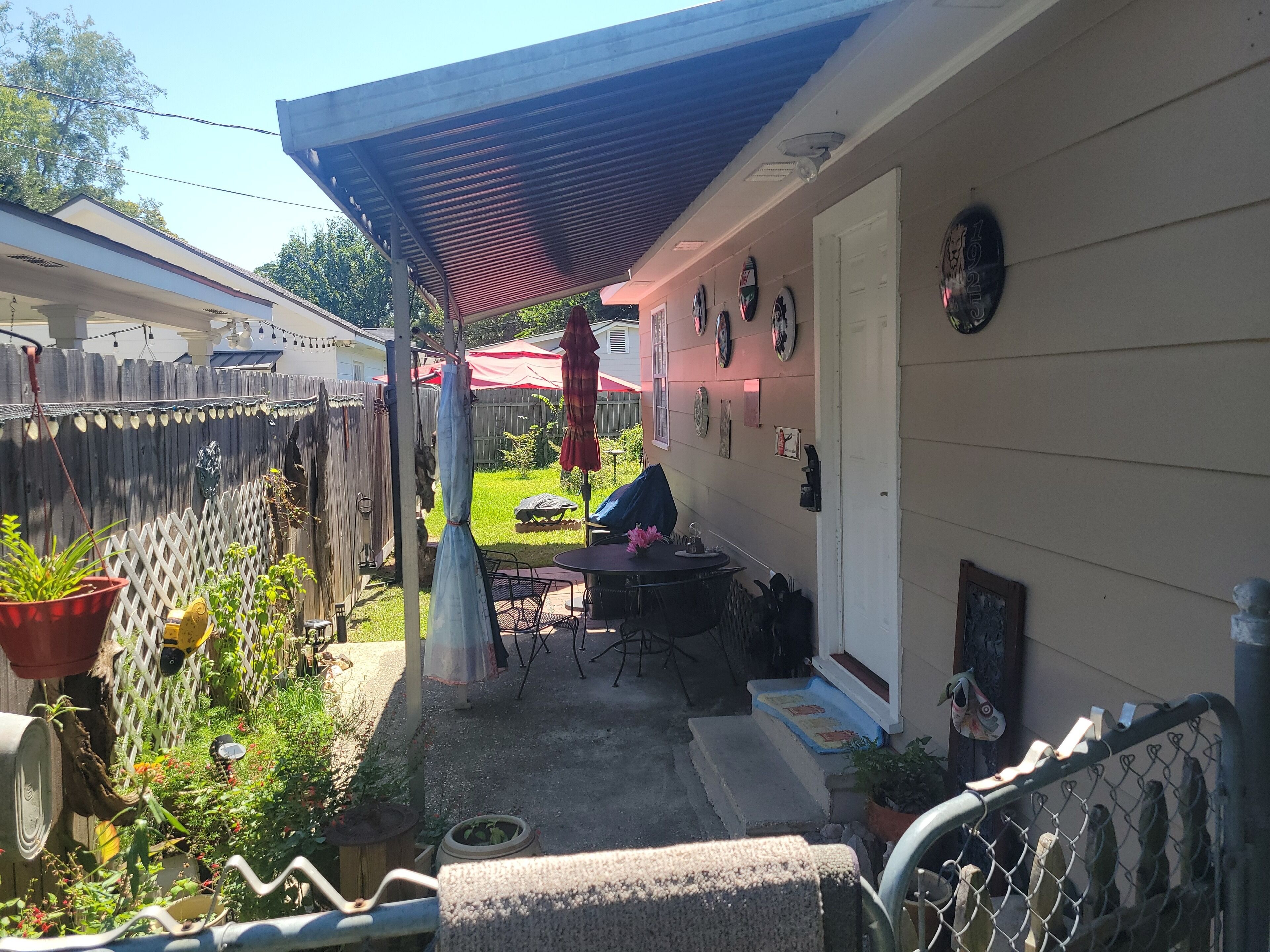 A covered patio area with a table and chairs, surrounded by potted plants and a wooden fence.
