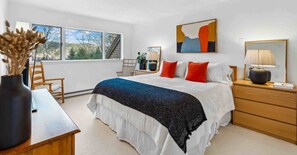 A bedroom with a bed, bedside table, lamp, and a window with a view of trees.